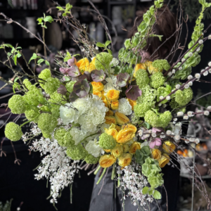 Duży, artystyczny bukiet w stylu ogrodowym trzymany przez florystkę. Widoczne żółte róże ogrodowe w gałązkowej odmianie, białe i limonkowe goździki, zielone kule kaliny, ciemierniki oraz surowe, wiosenne gałęzie i bazie. Domin ujące kolory: żółty, biały i limonkowy.